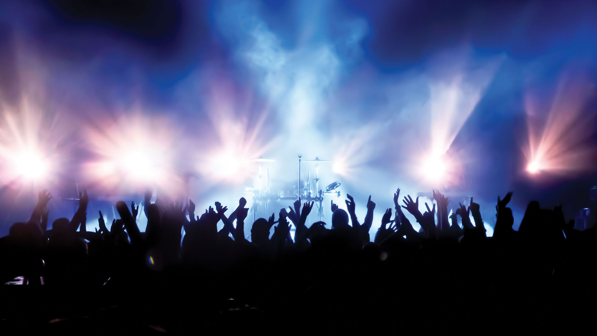 A live band on a concert stage with lights and a cheering audience silhouetted in the foreground