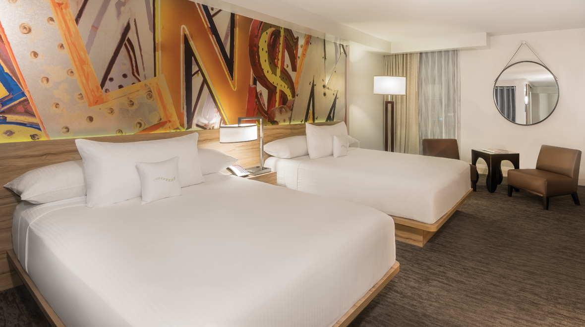 Hotel room with two beds, colorful mural, chairs, and a round mirror.