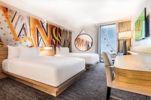 View Into A Linq Las Vegas Hotel Room, Showing A Bed, Nightstand, Mounted TV Above Dresser And A Seating Area Next To An Open Window