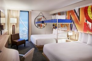Photo Of Hotel Room Showing Two Queen Beds, A Bunk Bed, A Chair And Desk Area, Mounted TV And Hanging Mirror At The Linq Las Vegas